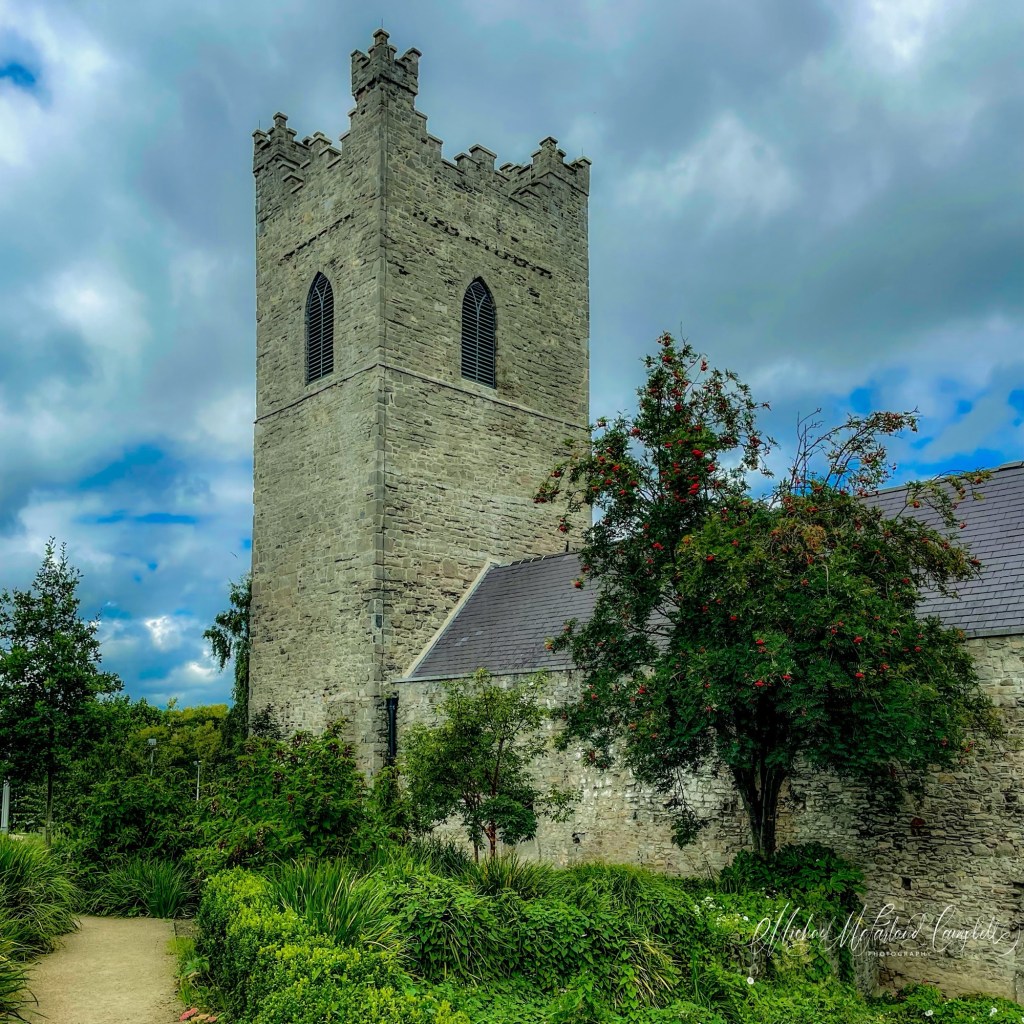 Medieval stone bell tower of St Audoen’s Church rising above green gardens beneath a cloudy blue sky.