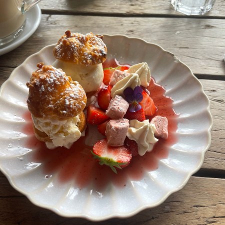 Two cream-filled profiteroles served with sliced strawberries, pink marshmallow pieces, piped cream, strawberry sauce and a purple edible flower on a white scalloped plate.